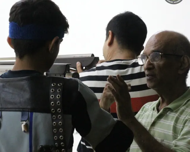 A rifle shooter in position, watched by a senior coach